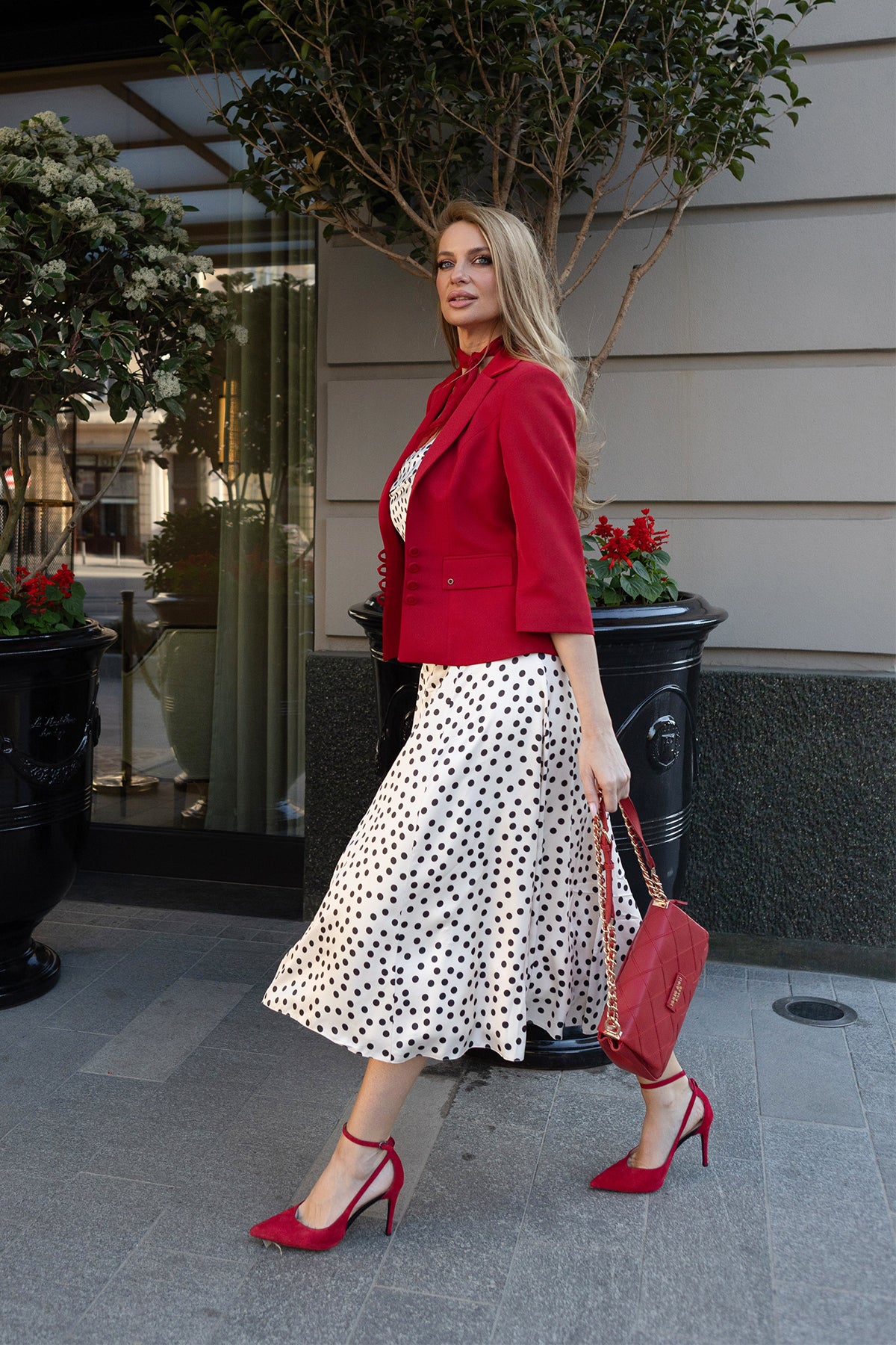 Woman walking on sidewalk wearing a red blazer, white polka dot dress, red heels, and carrying a red handbag.