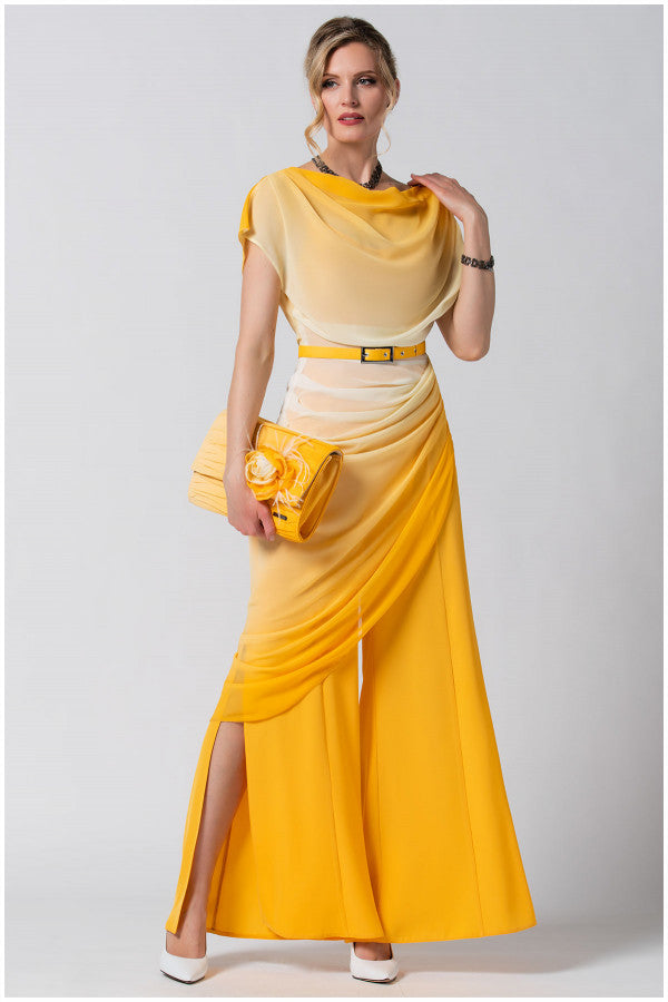 Woman wearing a flowing yellow and beige dress with side slits holding a matching yellow clutch against a plain background.