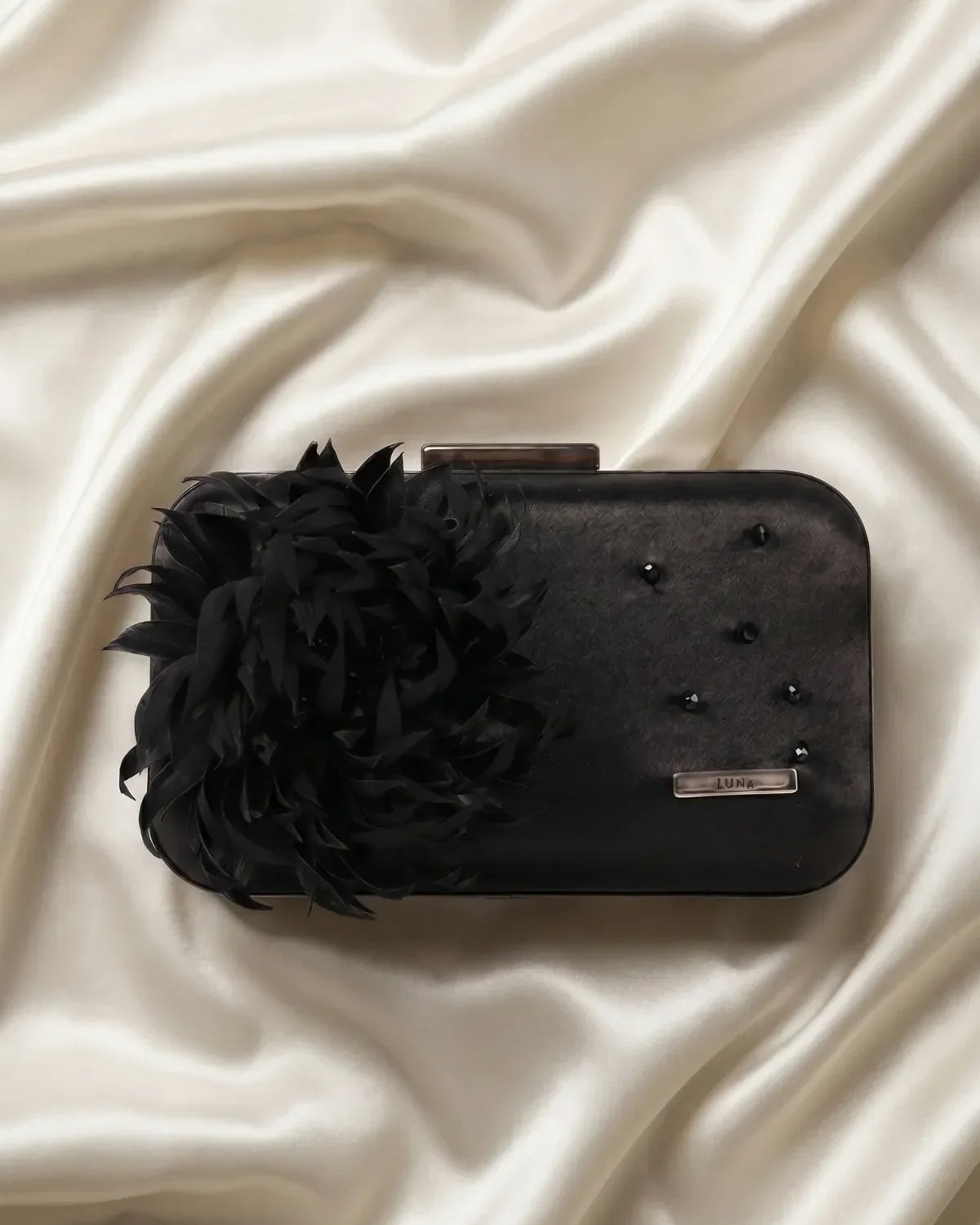 Structured black evening clutch with feather flower detail and subtle beadwork on a dark formal surface