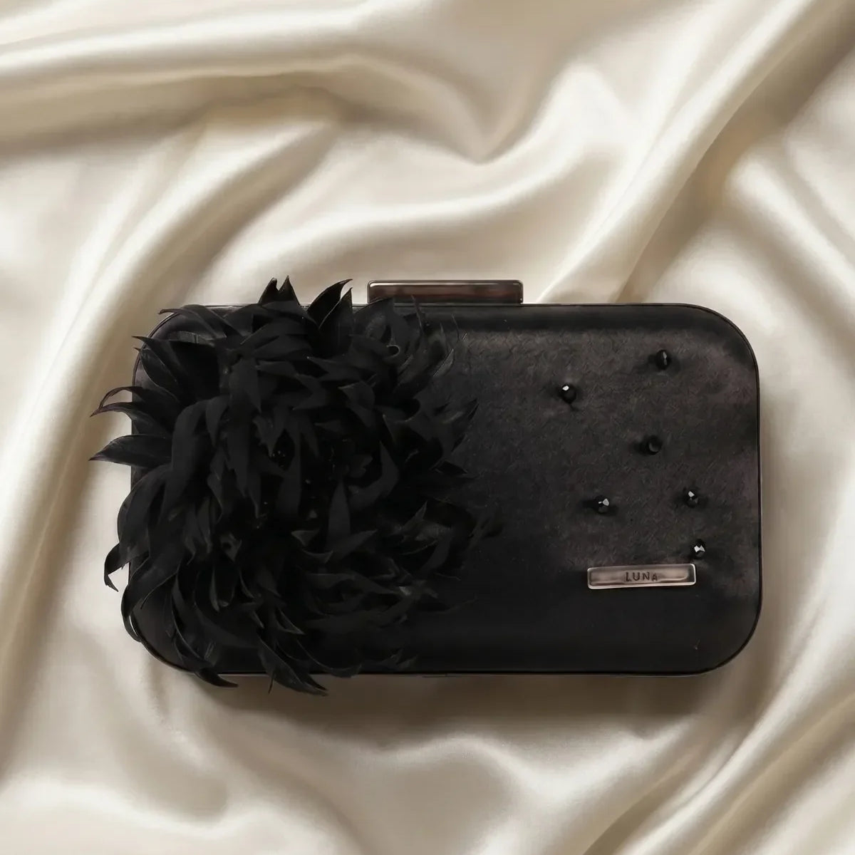 Black structured evening clutch with feather flower detail and removable chain strap on a neutral linen surface