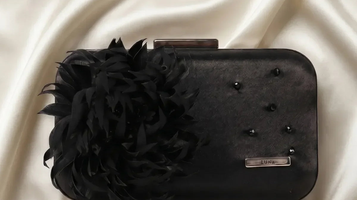 Black structured evening clutch with feather flower detail and removable chain strap on a neutral linen surface