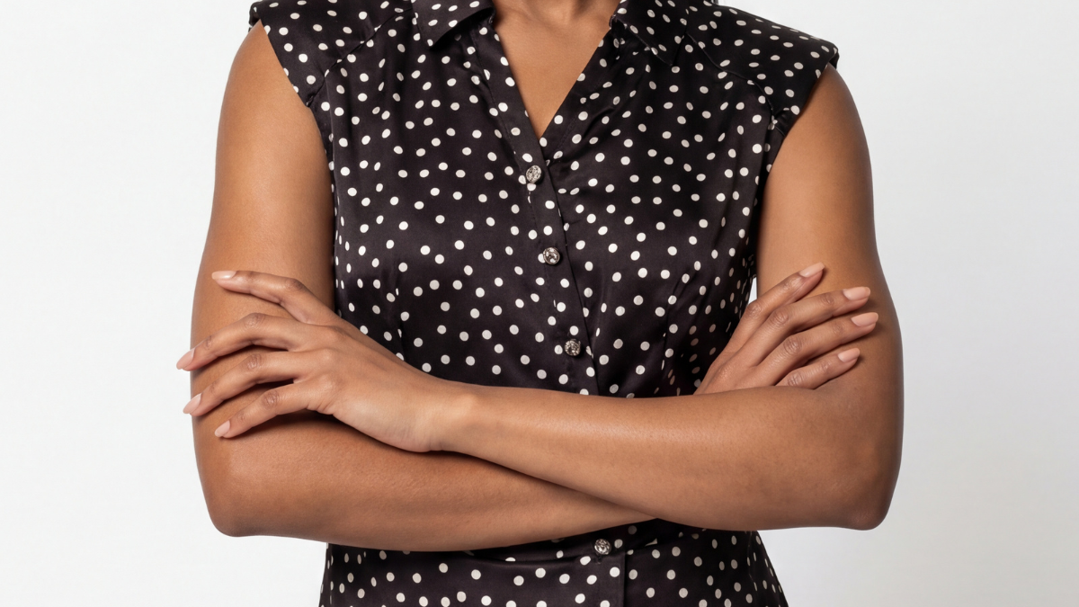 Black polka dot satin sleeveless V-neck blouse with diagonal button detail on a neutral studio background