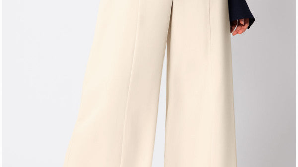 Ivory high-waisted wide-leg pants with a deep waistband, a metal buckle tab, a pressed center crease, side pockets, and a full straight leg to the floor, worn with a navy sheer blouse.
