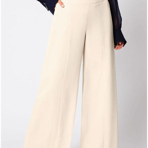 Ivory high-waisted wide-leg pants with a deep waistband, a metal buckle tab, a pressed center crease, side pockets, and a full straight leg to the floor, worn with a navy sheer blouse.
