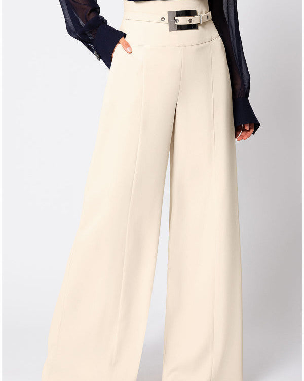 Ivory high-waisted wide-leg pants with a deep waistband, a metal buckle tab, a pressed center crease, side pockets, and a full straight leg to the floor, worn with a navy sheer blouse.