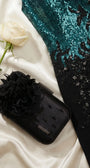 Black evening clutch beside a teal sequin gown and a white rose on satin