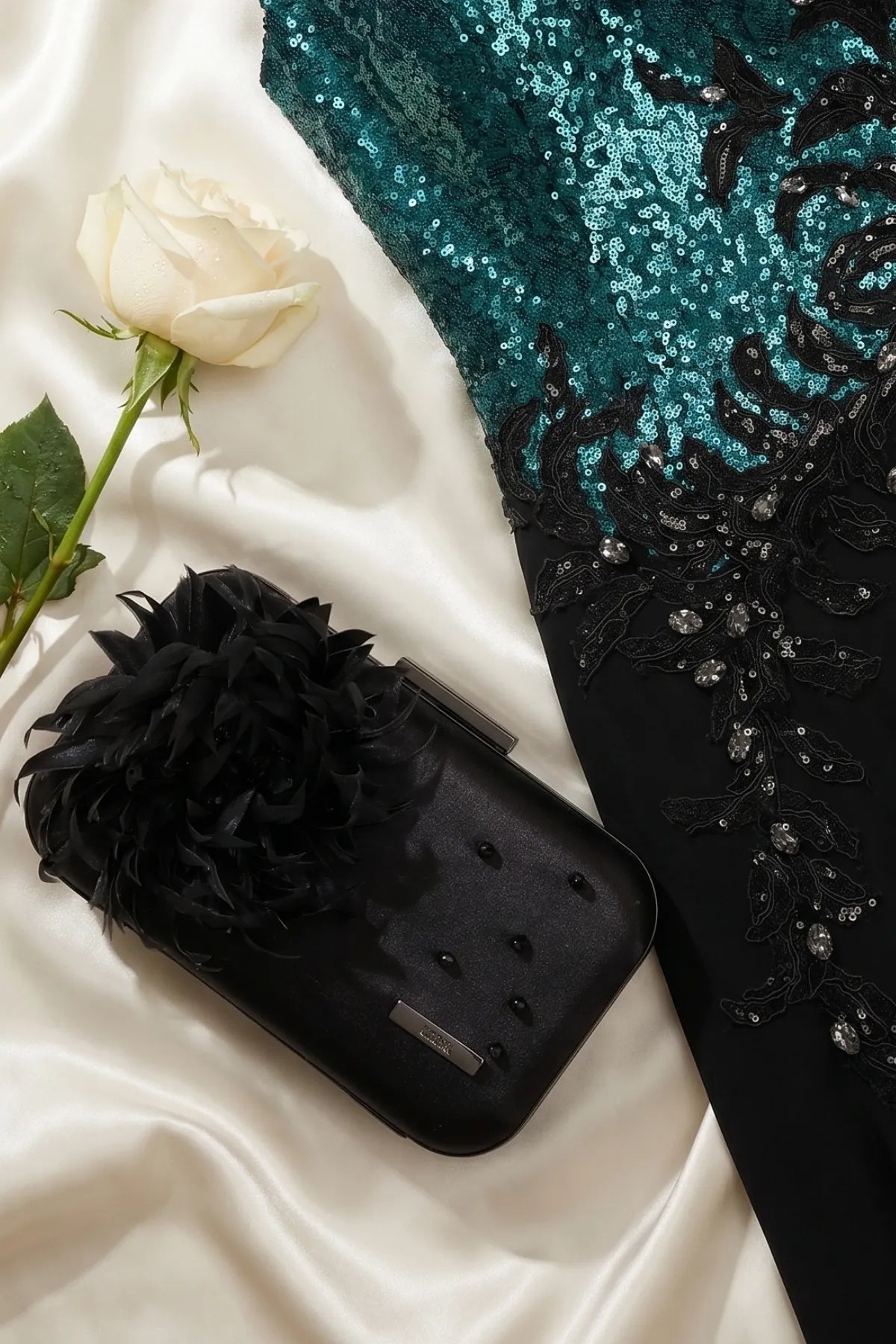 Black evening clutch beside a teal sequin gown and a white rose on satin