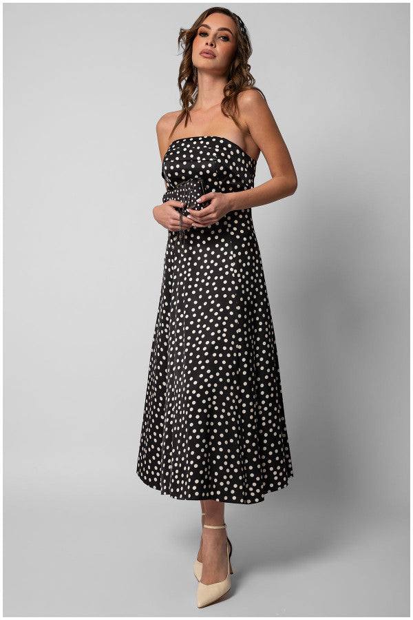 Woman wearing a black elegant midi dress satin strapless midi dress with white polka dots and beige heels holding a small clutch bag.