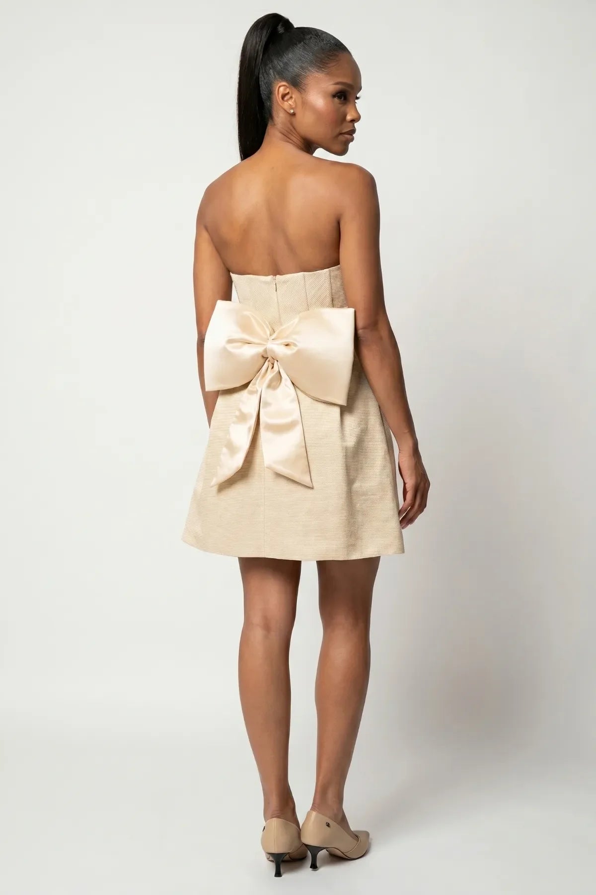 Angled back view of Melisa beige strapless mini dress with large bow and flared skirt.