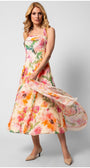 Woman wearing a flowing floral dress with pink, orange, and green hues on a light background, standing against gray wall.
