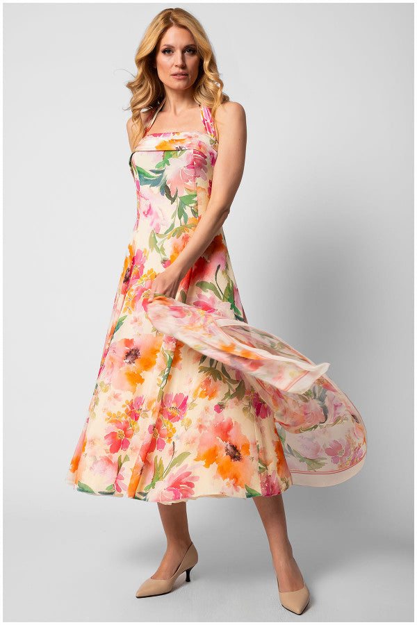 Woman wearing a flowing floral dress with pink, orange, and green hues on a light background, standing against gray wall.