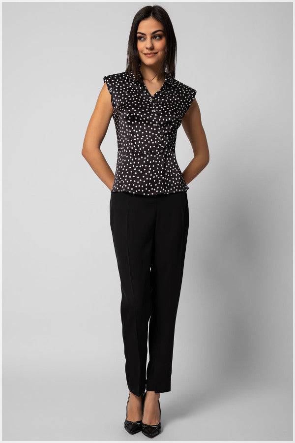 Woman wearing a sleeveless black satin elegant blouse with white polka dots and decorative diagonal buttons paired with tailored black pants.