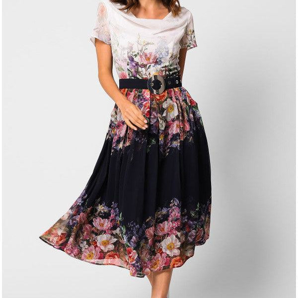 Woman wearing elegant midi dress Julia Dress with floral pattern, black belt, flowing midi length, and white pointed shoes on white background.