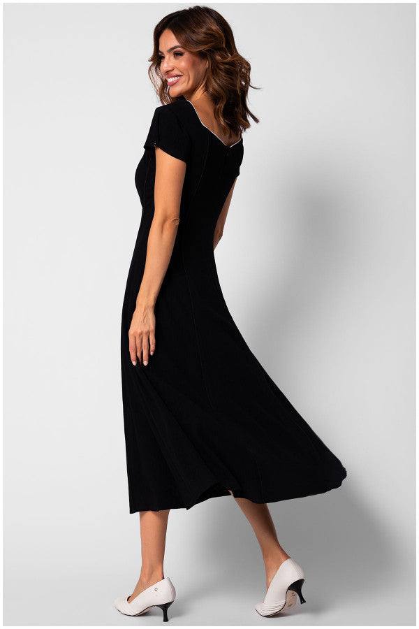 Woman wearing a black elegant midi dress Rina with short sleeves and square neckline, paired with white heels, smiling and posing.