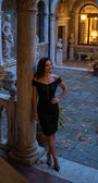 Woman wearing the black ruched off-shoulder cocktail midi in a grand stone courtyard at dusk with statues and lantern light