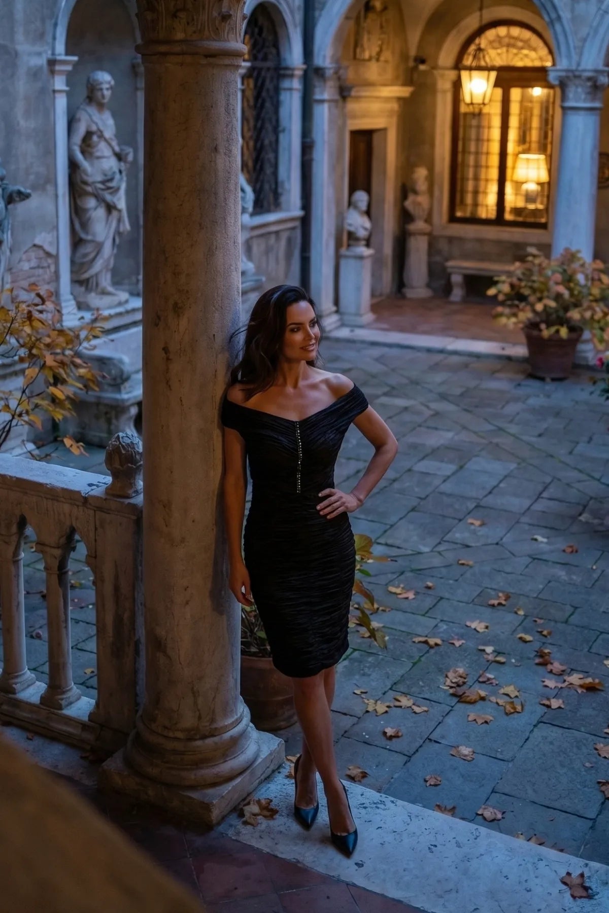 Woman wearing the black ruched off-shoulder cocktail midi in a grand stone courtyard at dusk with statues and lantern light