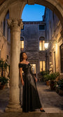 Woman in an anthracite metallic off-shoulder maxi gown in a stone courtyard at dusk lit by a warm lantern