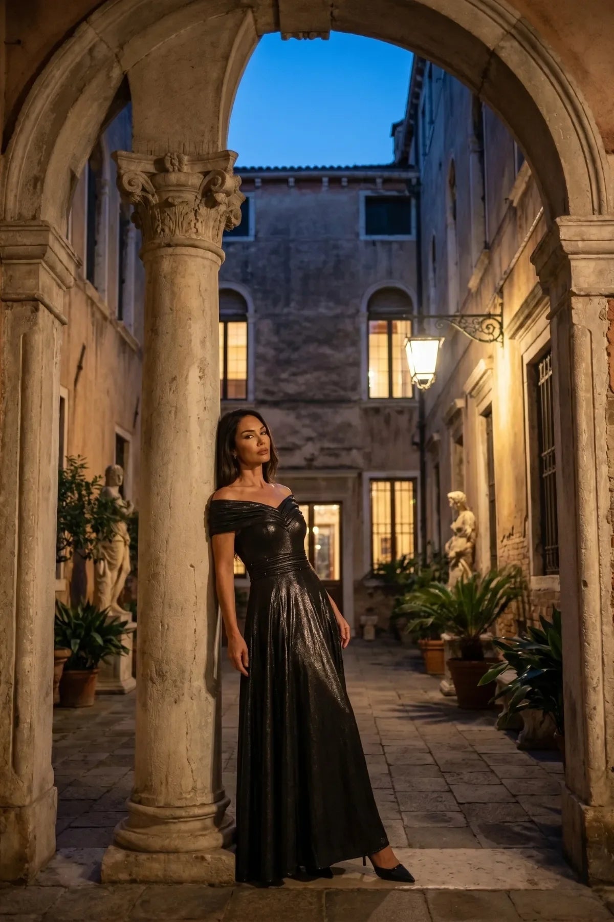 Woman in an anthracite metallic off-shoulder maxi gown in a stone courtyard at dusk lit by a warm lantern