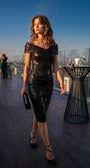 Model wearing the black cocktail dress at an evening rooftop event with city skyline at sunset.