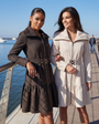 Beige Cotton Short Tailored Coat, Iris. Short coat. Worn zipped with the clasp belt fastened, beside the brown Iris coat, on a marina boardwalk.