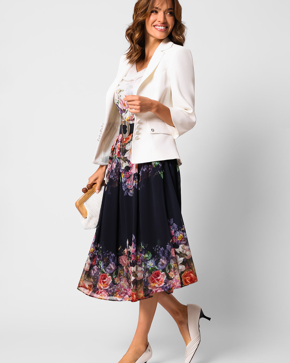 Woman wearing a white blazer and floral midi dress holding a small woven handbag against a plain background.