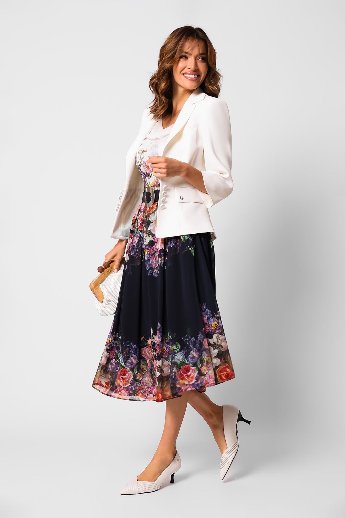 Woman wearing a white blazer and floral midi dress holding a small woven handbag against a plain background.
