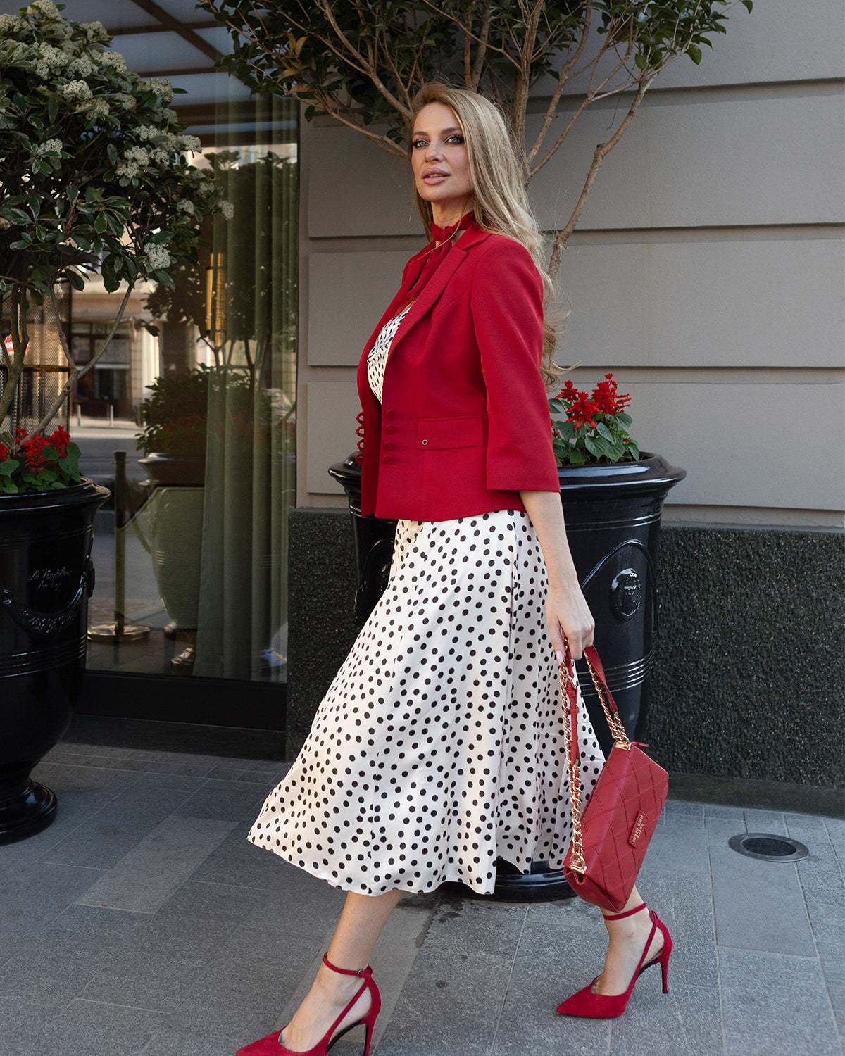 Woman walking on sidewalk wearing a red blazer, white polka dot dress, red heels, and carrying a red handbag.