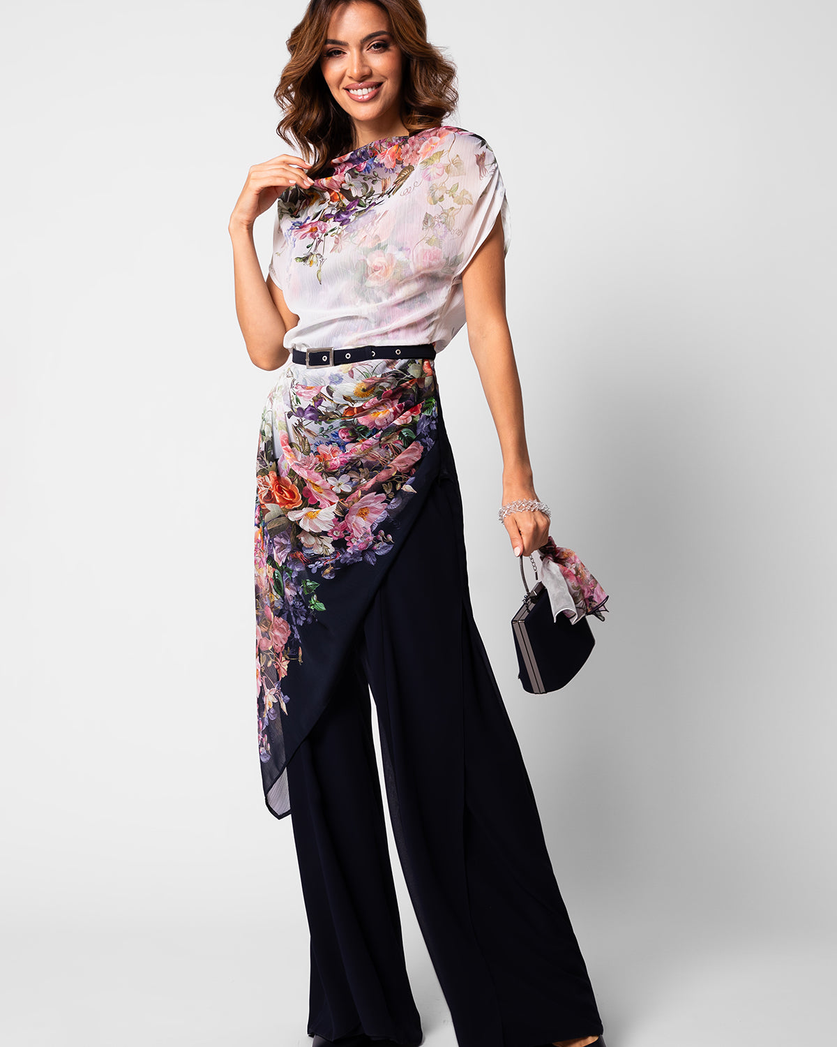 Smiling woman wearing floral sheer top and wrap scarf over wide-leg black pants holding a small handbag.