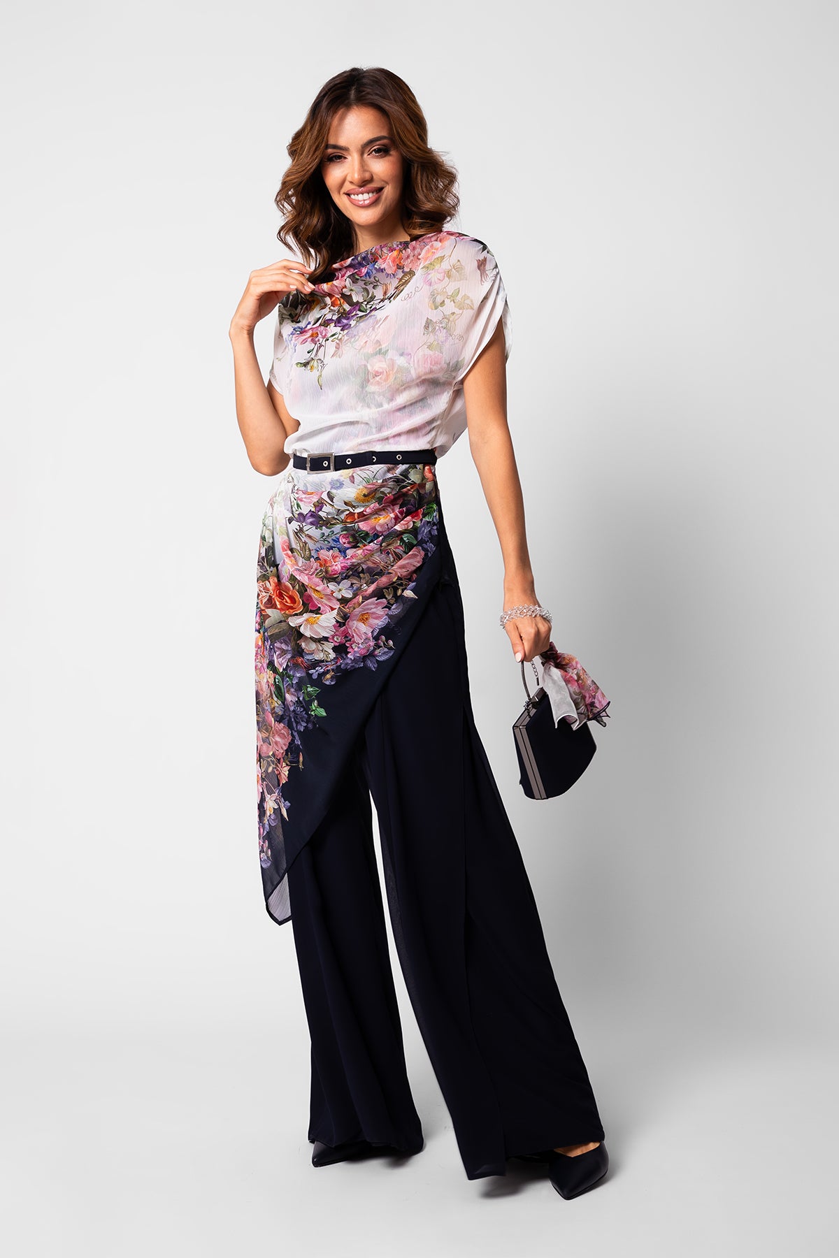 Smiling woman wearing floral sheer top and wrap scarf over wide-leg black pants holding a small handbag.