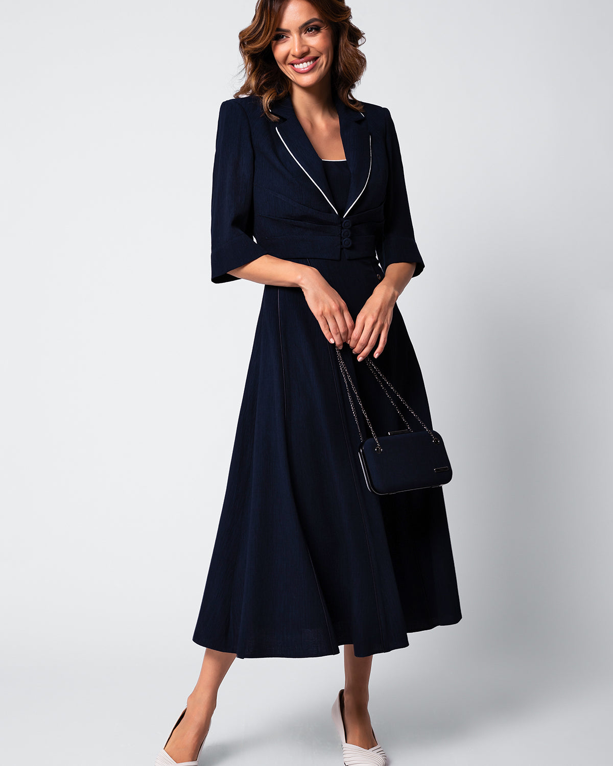 Woman in a navy midi dress with three-quarter sleeves holding a black handbag and wearing white pointed shoes.