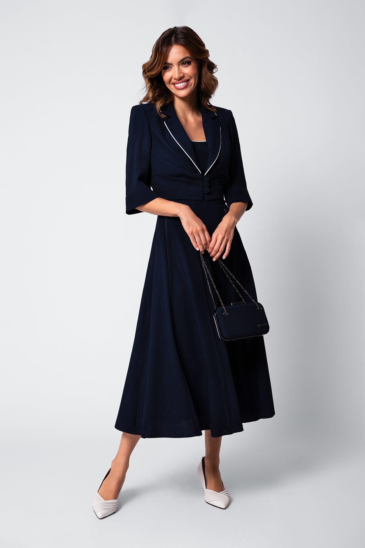 Woman in a navy midi dress with three-quarter sleeves holding a black handbag and wearing white pointed shoes.