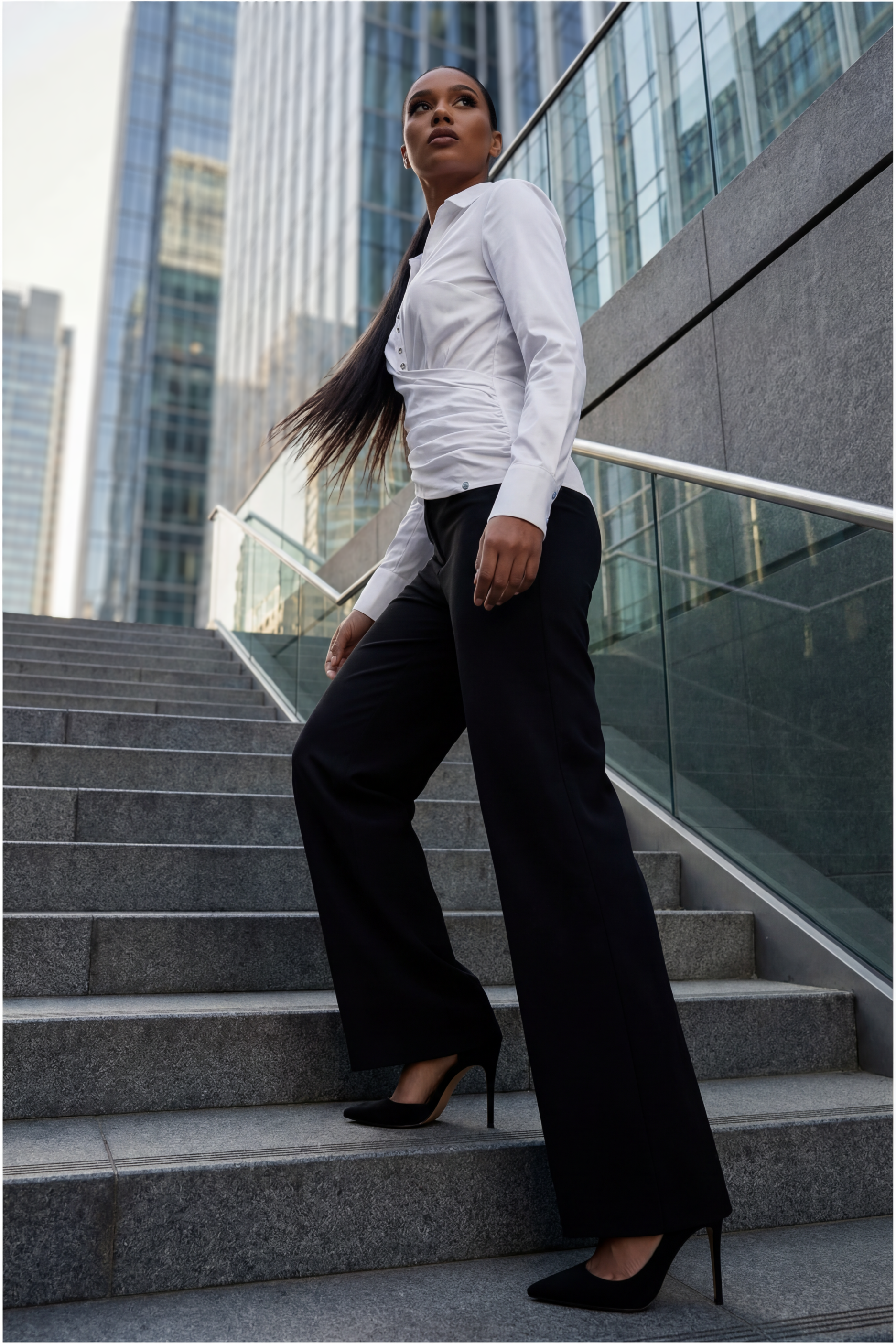Woman in a white blouse and black pants standing on steps with modern buildings in the background