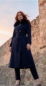 Woman wearing a long navy coat with fur collar standing in front of stone ruins.