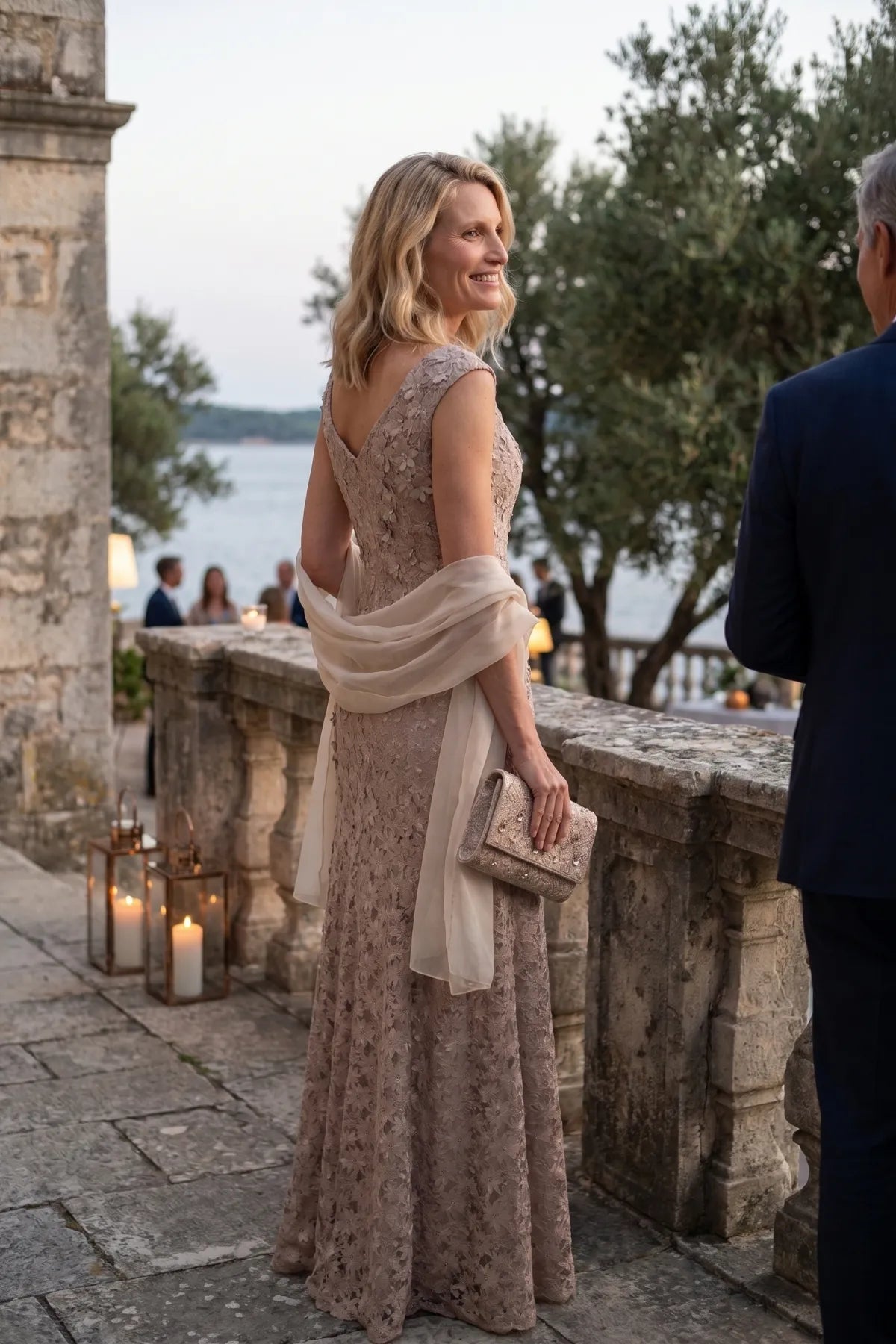 A woman in the beige lace gown looks back over her shoulder on a stone terrace by candlelight and water