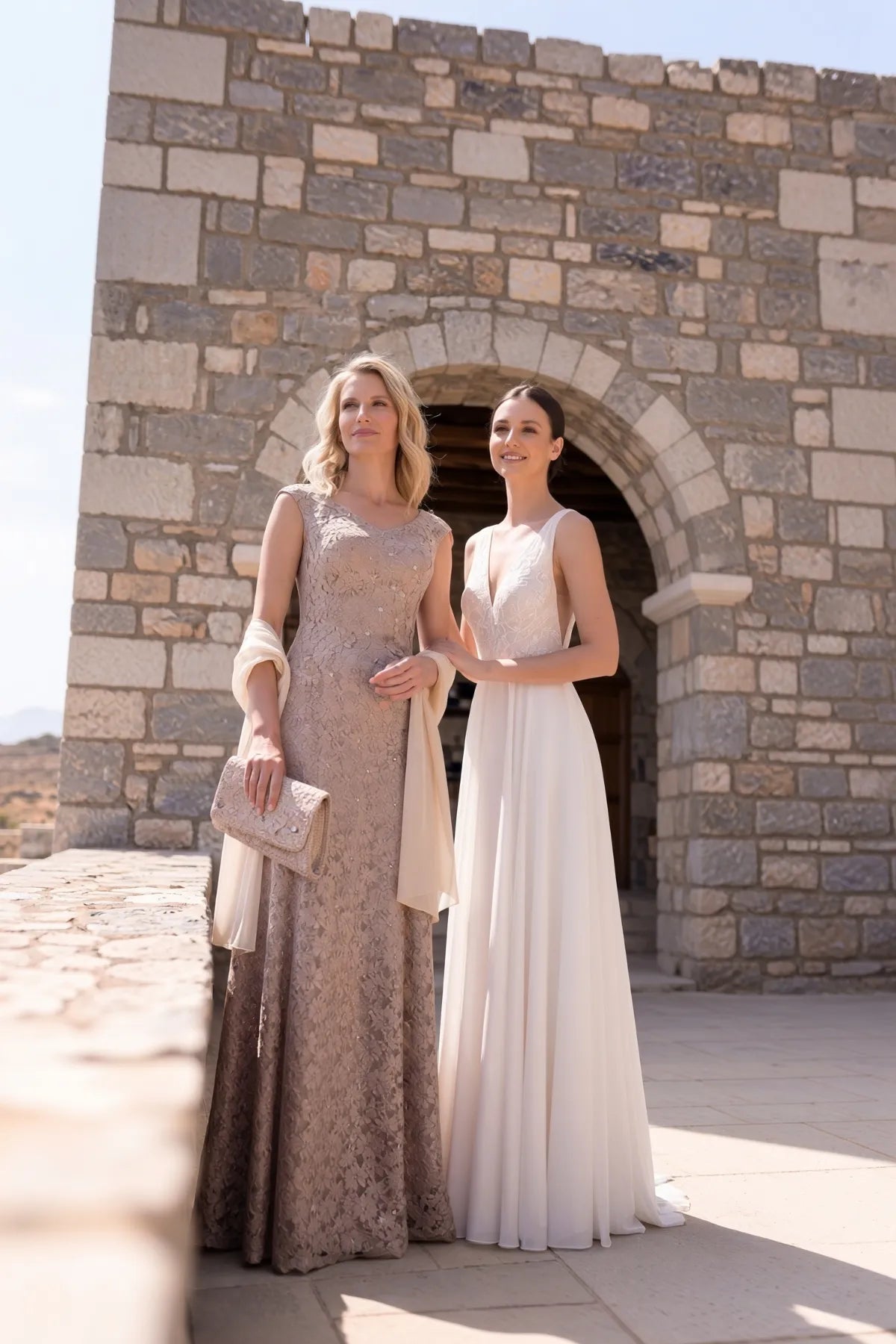 A woman in the beige lace gown stands beside a bride at a stone archway venue for a destination wedding