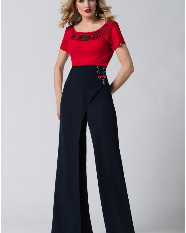 Woman with wavy blonde hair wearing a red top and high-waisted wide-leg navy pants against a gray background.