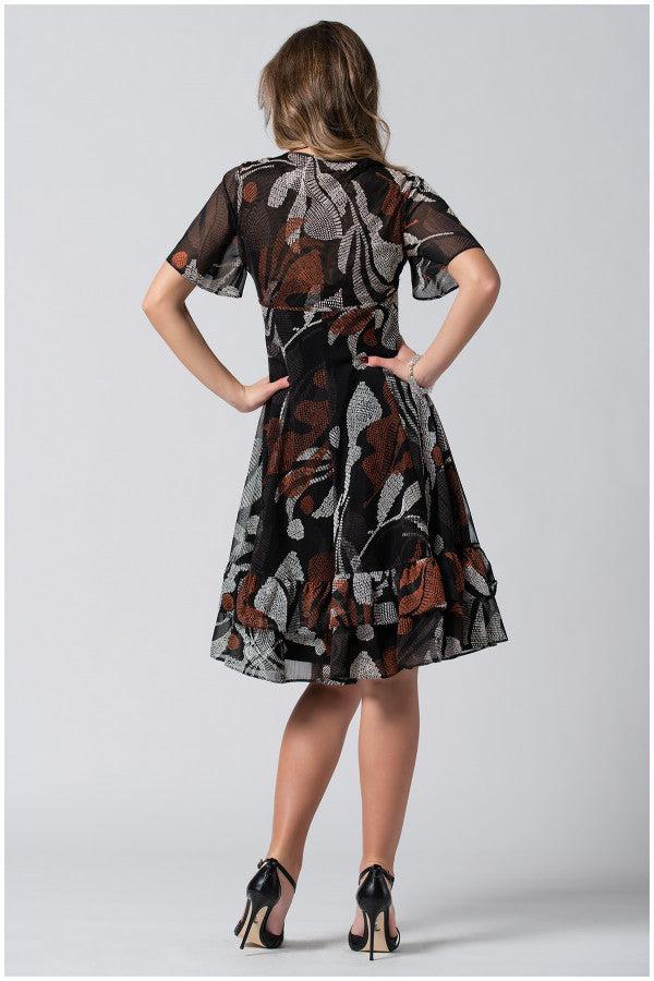 Woman wearing a black and brown patterned short wide dress with flounces, standing with hands on hips, back view.
