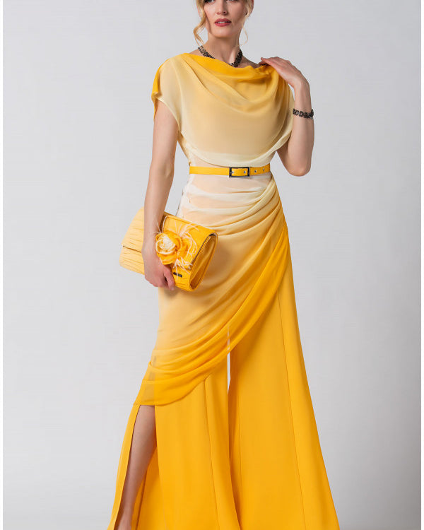 Woman wearing a flowing yellow and beige dress with side slits holding a matching yellow clutch against a plain background.
