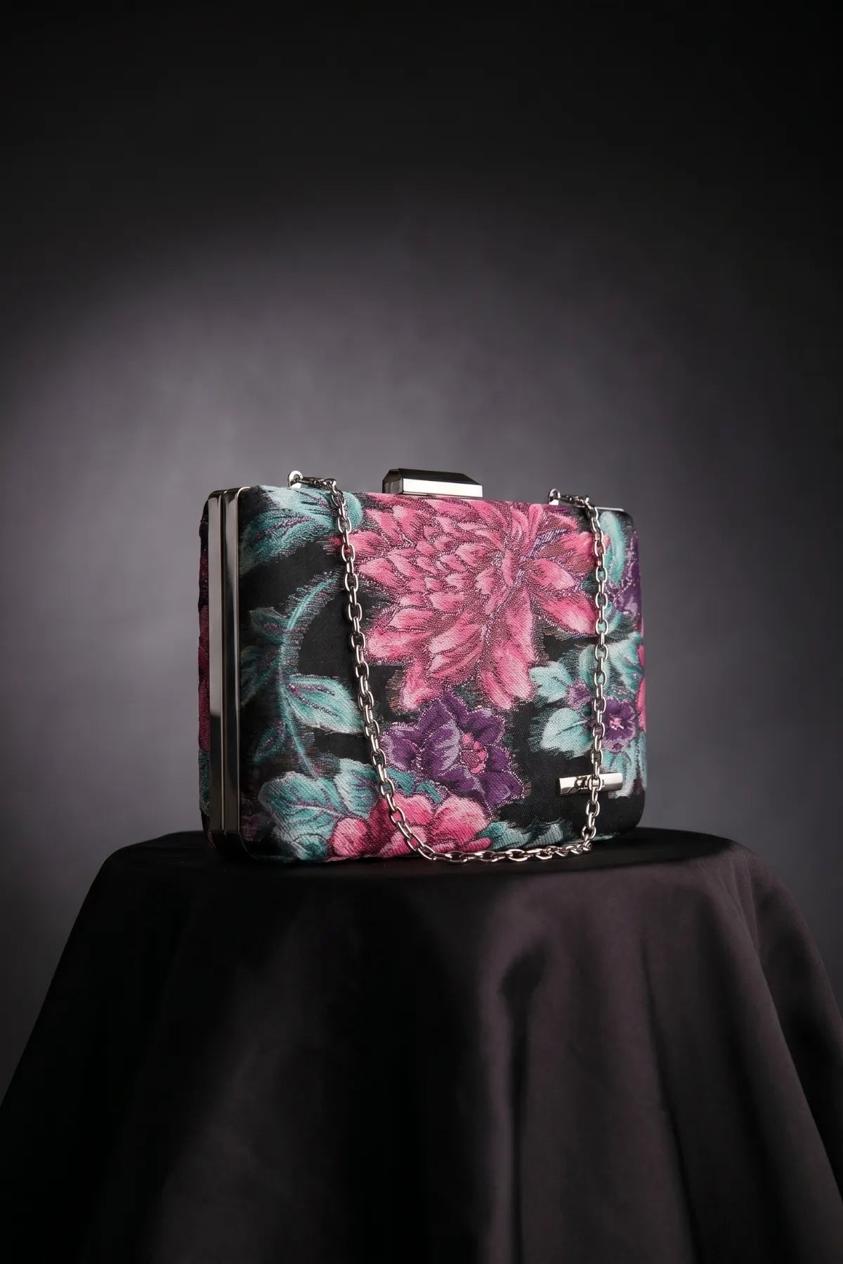 Regina floral clutch in cyclamen, purple, and teal on a dark surface with a detachable chain strap.