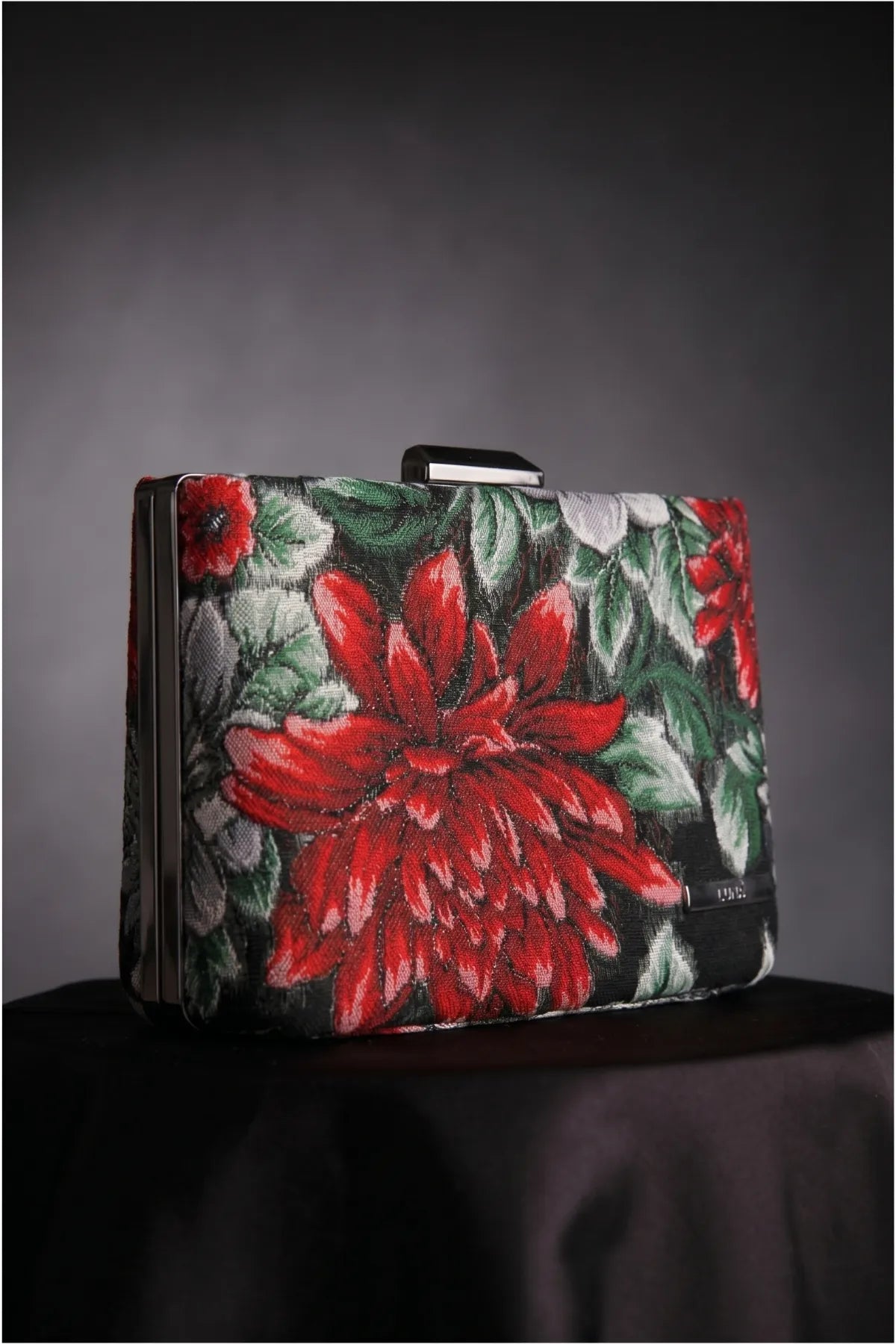 Regina red floral clutch on a dark draped surface highlighting the red floral motif and structured shape.