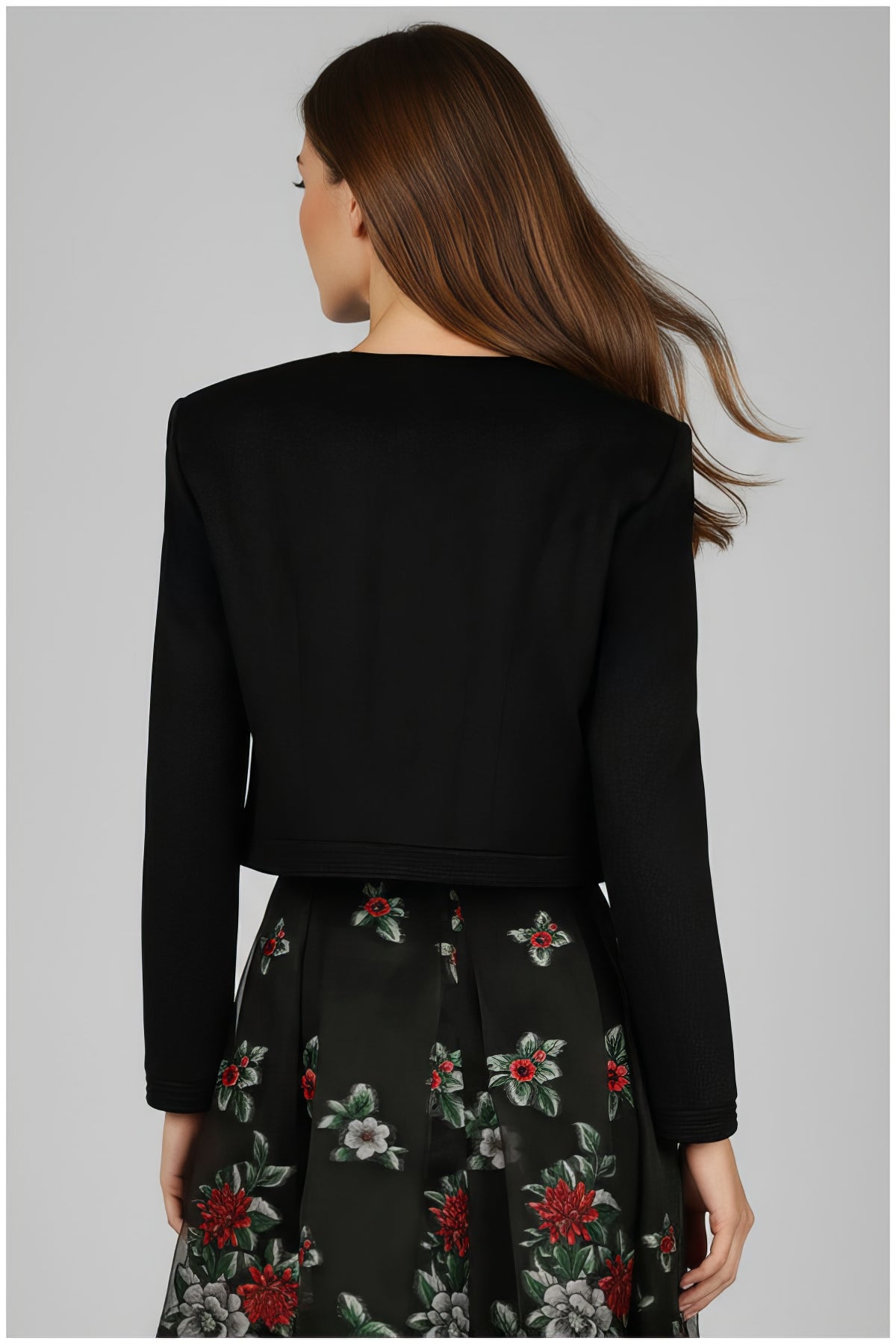 Back view of a woman wearing the Frida Cropped Zip Blazer in Black with a floral skirt against a gray background