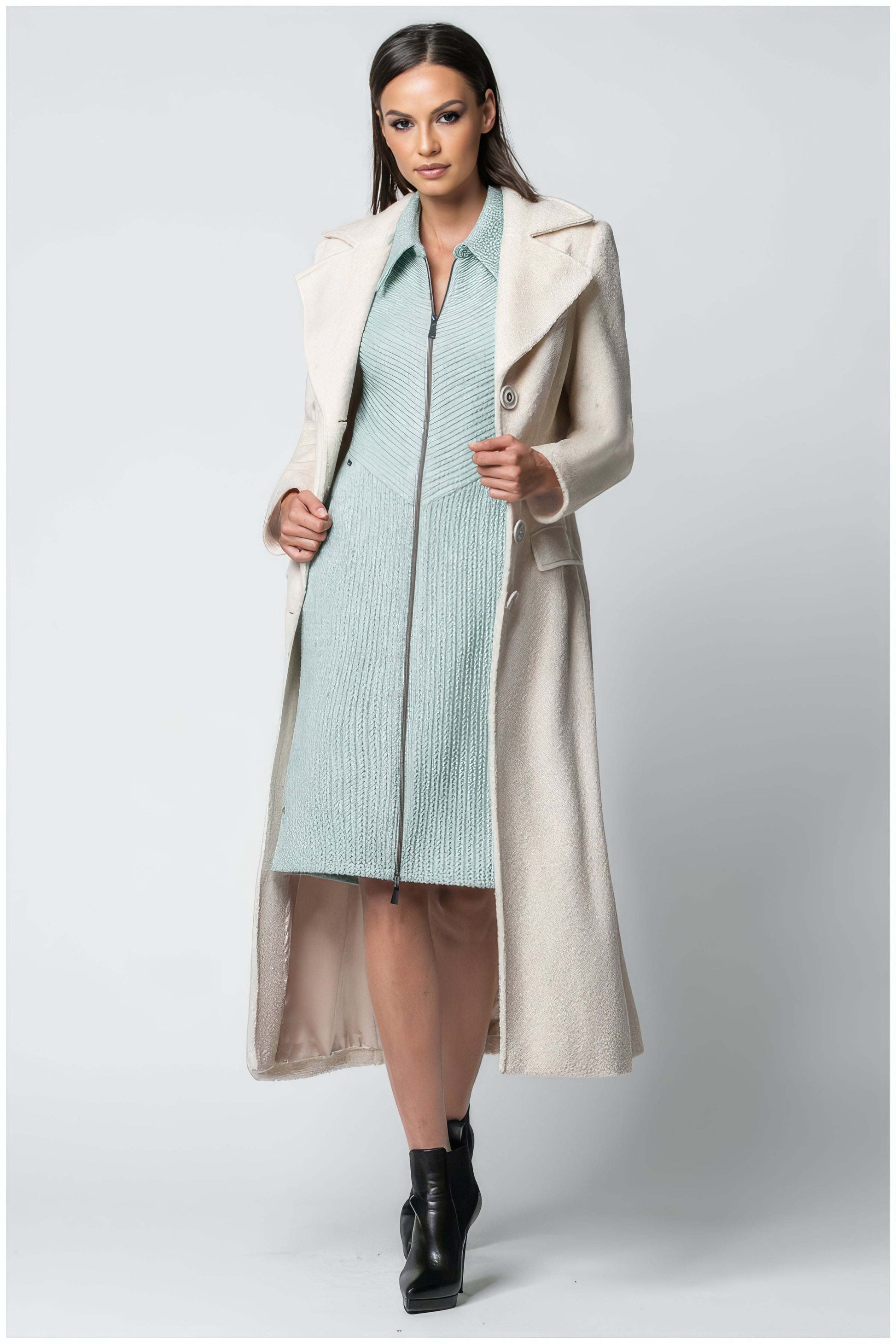 Woman wearing the Linea Belted Midi Coat in Ivory styled with a light blue dress and black ankle boots on white background