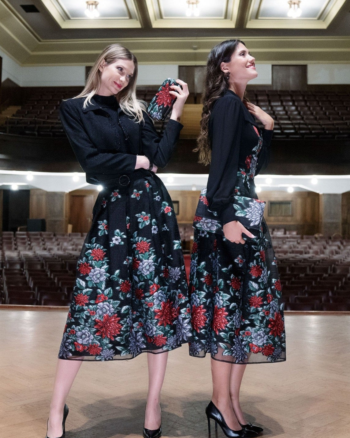 Two women wearing black floral dresses in an auditorium.