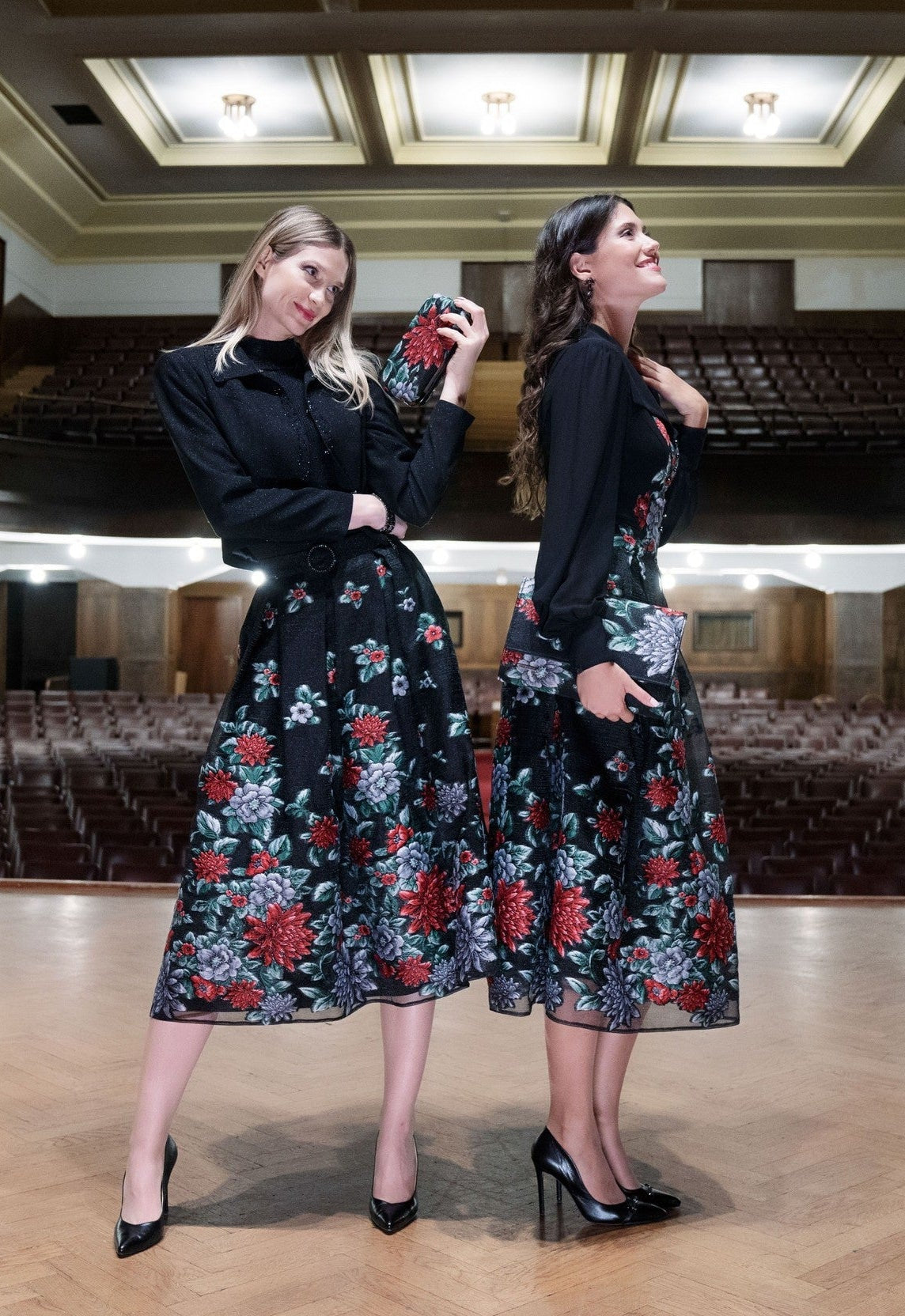 Two women wearing black floral dresses in an auditorium.