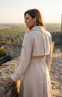 Beige Classic Long Coat, Kim. Back view on a fortress wall, showing the back storm flap, epaulettes, and buttoned sleeve strap.