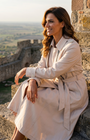 Beige Classic Long Coat, Kim. Worn closed with the belt tied, seated on stone fortress steps at golden hour.