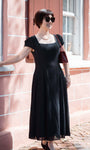 Woman wearing Rina Elegant Midi Dress in Black standing outdoors by a window in stylish sunglasses and pearl necklace