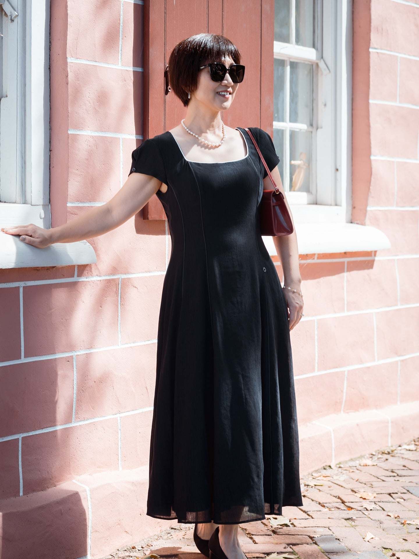 Woman wearing Rina Elegant Midi Dress in Black standing outdoors by a window in stylish sunglasses and pearl necklace