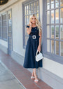 Woman wearing Greta Off-Shoulder Dress in Navy holding white handbag standing by window in elegant setting